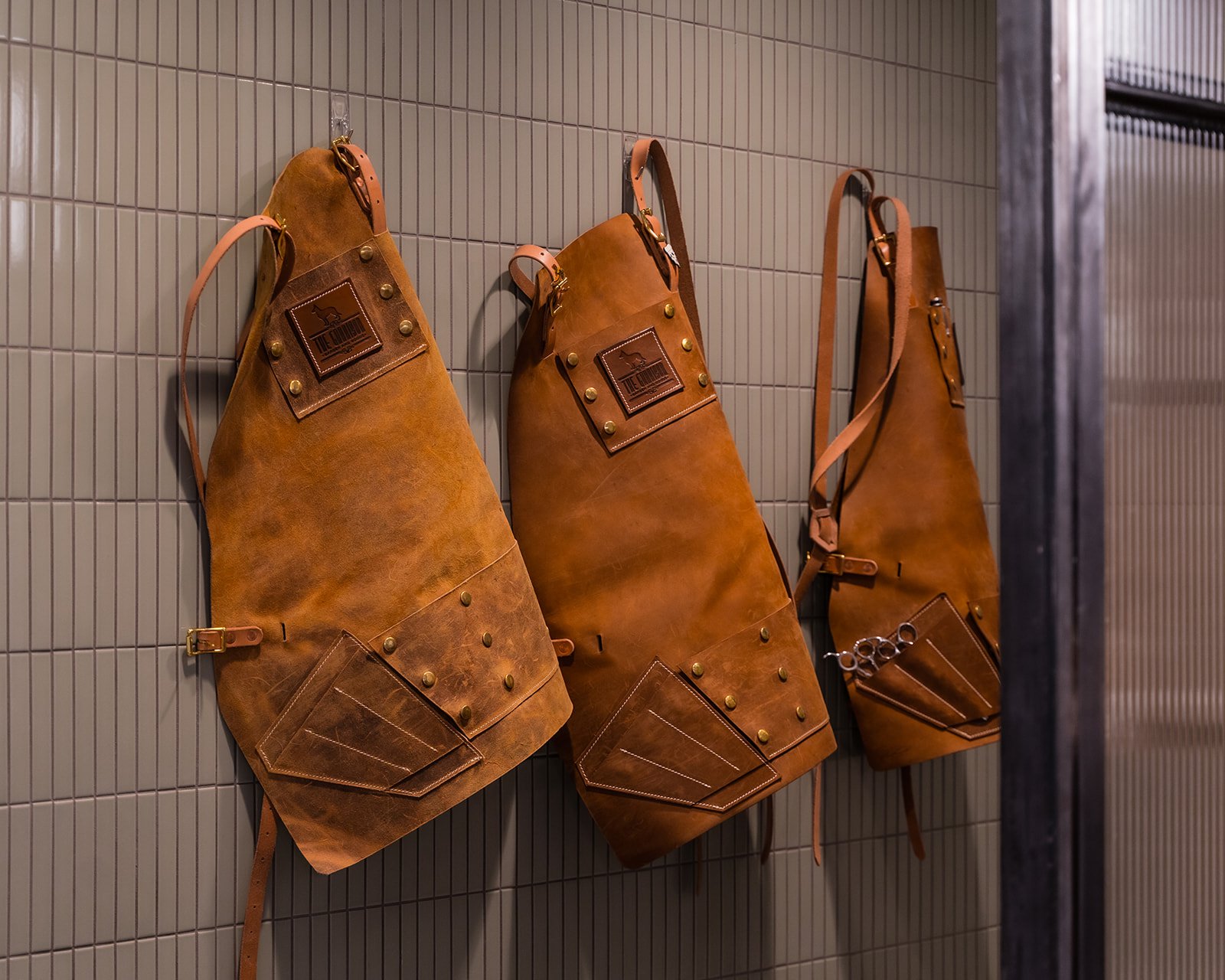Three leather barber aprons hanging on the tiled wall of the shop